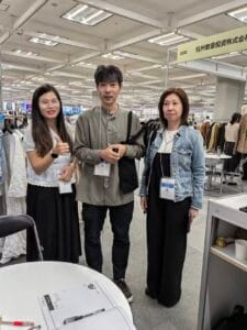 Three people stand together at an indoor trade show booth, smiling for the camera. A woman on the left gives a thumbs up. Clothing racks and tables are visible in the background.