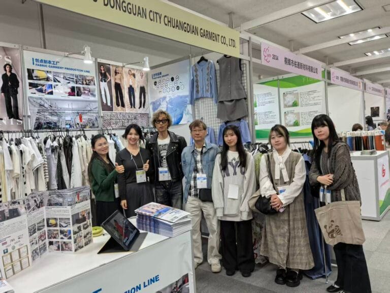 A group of six people stand in front of a trade show booth for Dongguan City Chuangtian Garment Co., Ltd., surrounded by clothing displays, brochures, and informational posters.