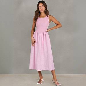 A woman stands in front of a gray background wearing a sleeveless, light pink midi dress and white heeled sandals. She has long, wavy brown hair and poses with one hand on her hip. A woman stands in front of a gray background wearing a sleeveless, light pink midi dress and white heeled sandals. She has long, wavy brown hair and poses with one hand on her hip.