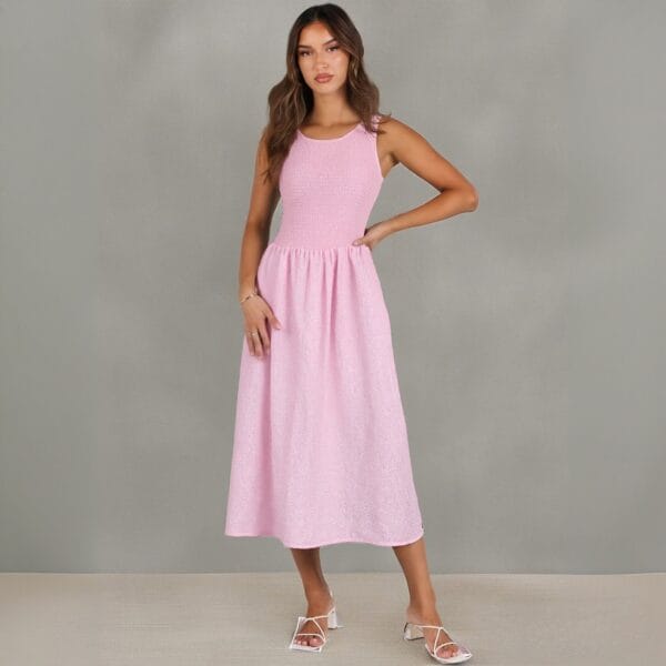 A woman stands in front of a gray background wearing a sleeveless, light pink midi dress and white heeled sandals. She has long, wavy brown hair and poses with one hand on her hip. A woman stands in front of a gray background wearing a sleeveless, light pink midi dress and white heeled sandals. She has long, wavy brown hair and poses with one hand on her hip.